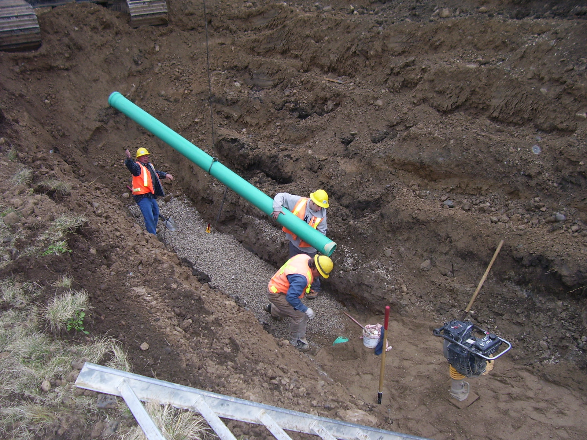 Workers lowering green sewer pipe into trench